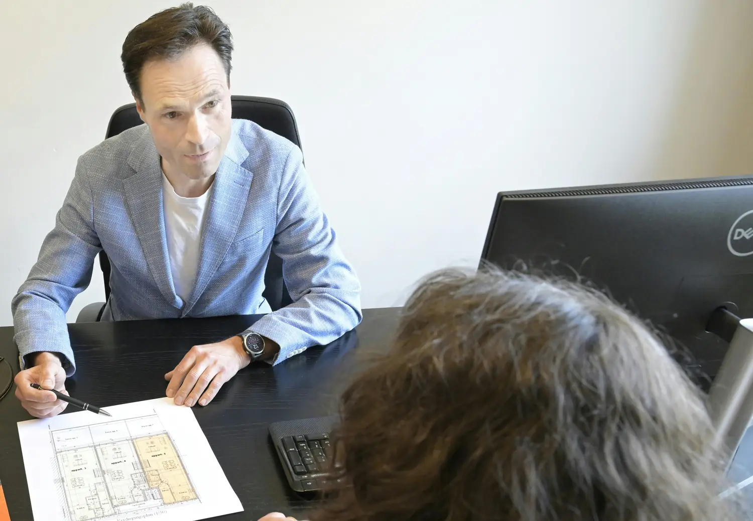 Across a desk, a man in a light blazer goes through a plan with a client.