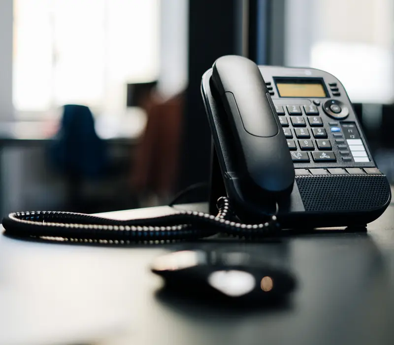 An office desk phone on a dark desk by a window.